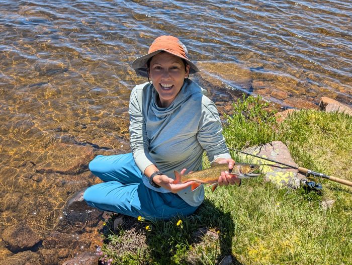 A woman holding a brook trout sitting at the edge of a lake