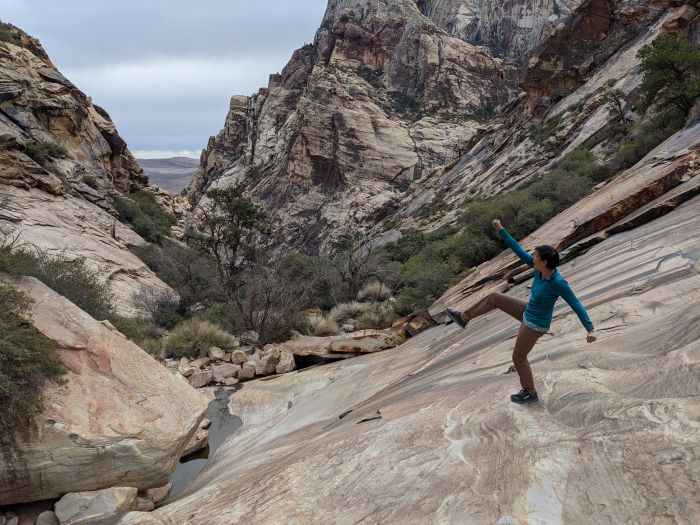 A woman standing on one leg with an arm in the air on a large flat rock with rocky hills in the background